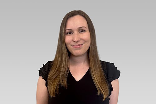 Woman with straight brown hair, wearing a black short-sleeve top, standing and smiling in front of a plain light gray background.