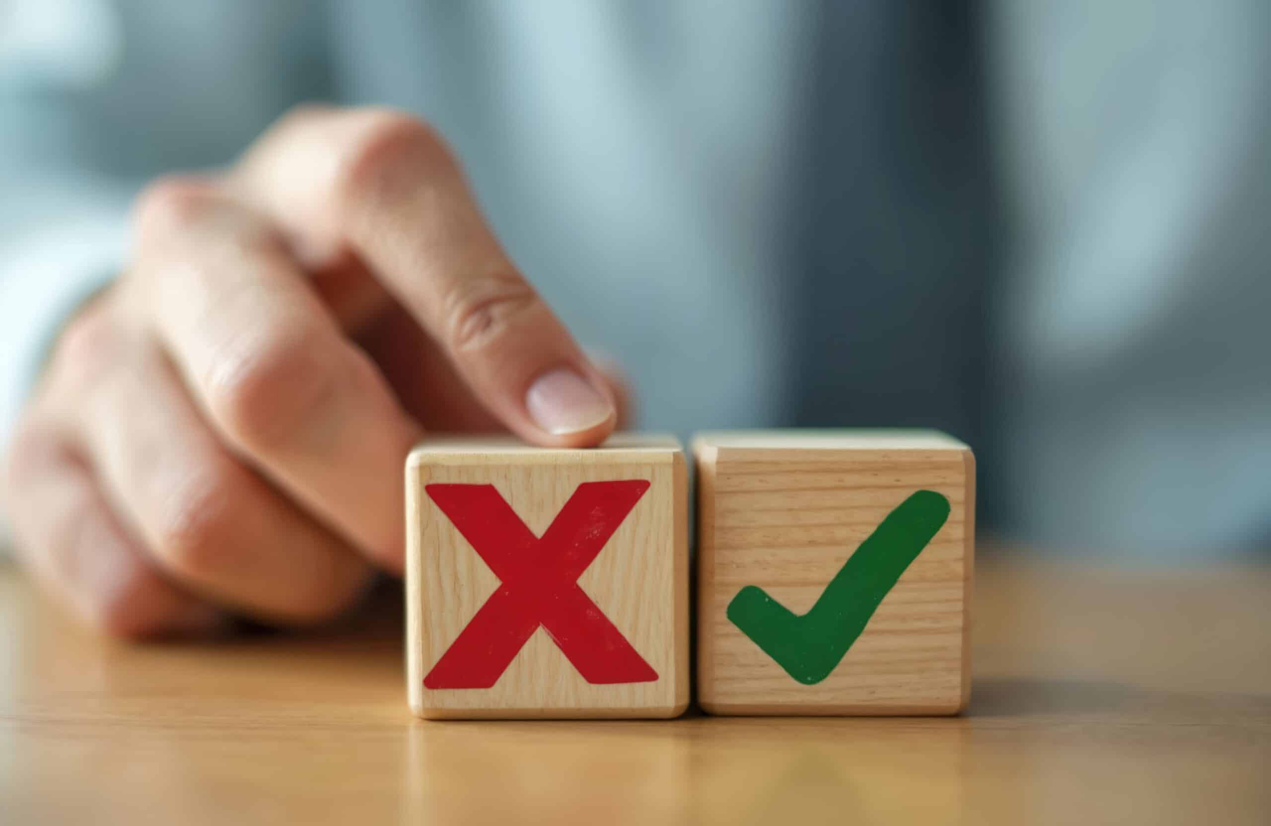 A hand points at two wooden blocks, one with a red X and the other with a green check mark, on a wooden surface.